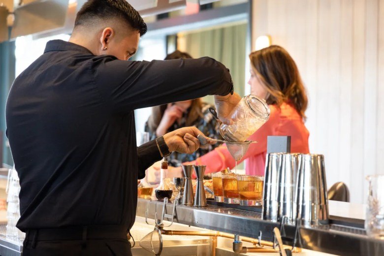 A bartender in a black shirt straining a drink into a glass at a bar counter.