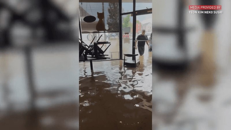 Person pushing murky brown floodwater across a commercial floor with overturned furniture visible.