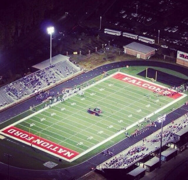 Aerial night view of a football stadium with green turf, blue track, and lit bleachers.