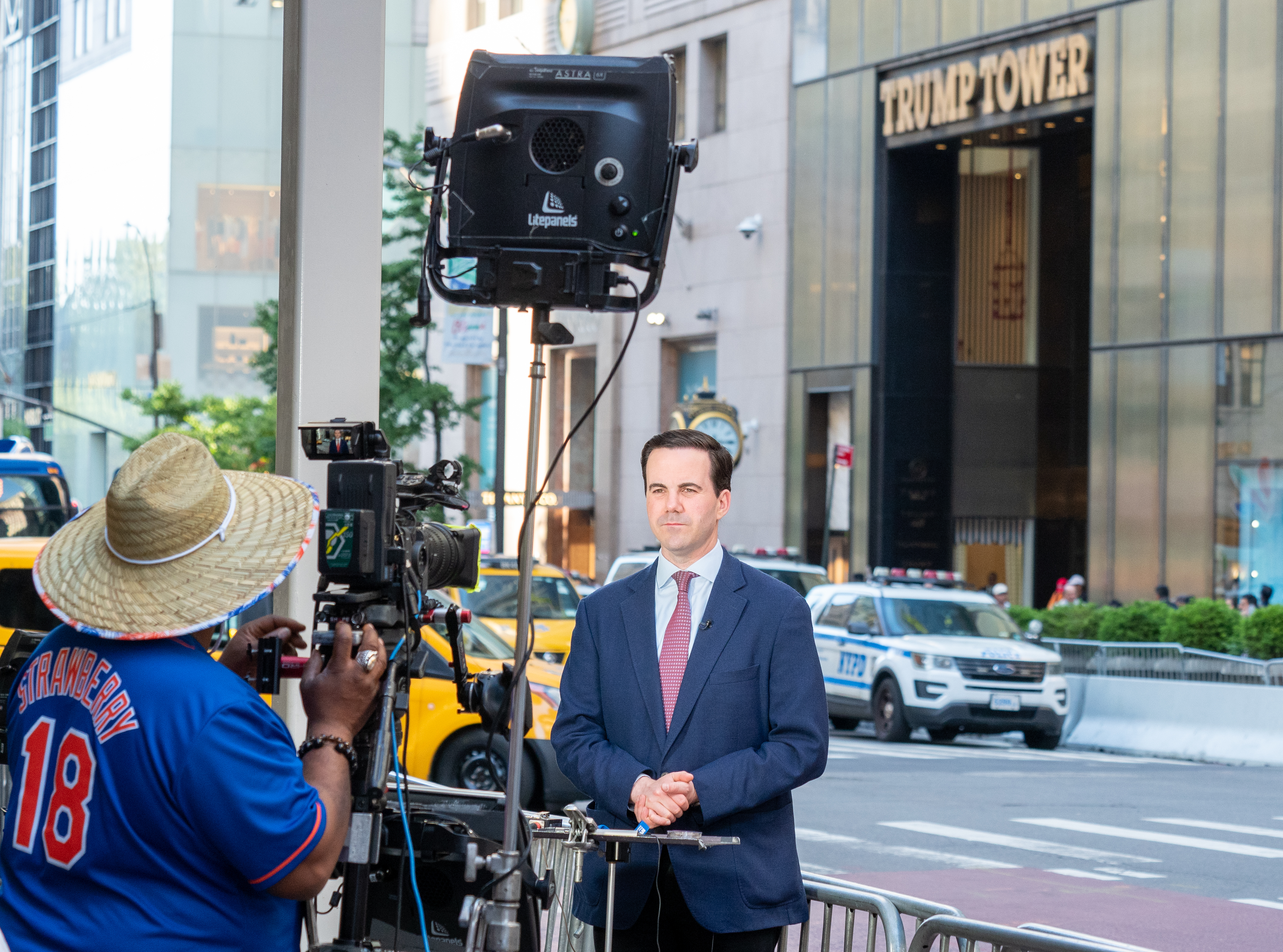 A man in a navy suit stands before a skyscraper as a camera operator films him.