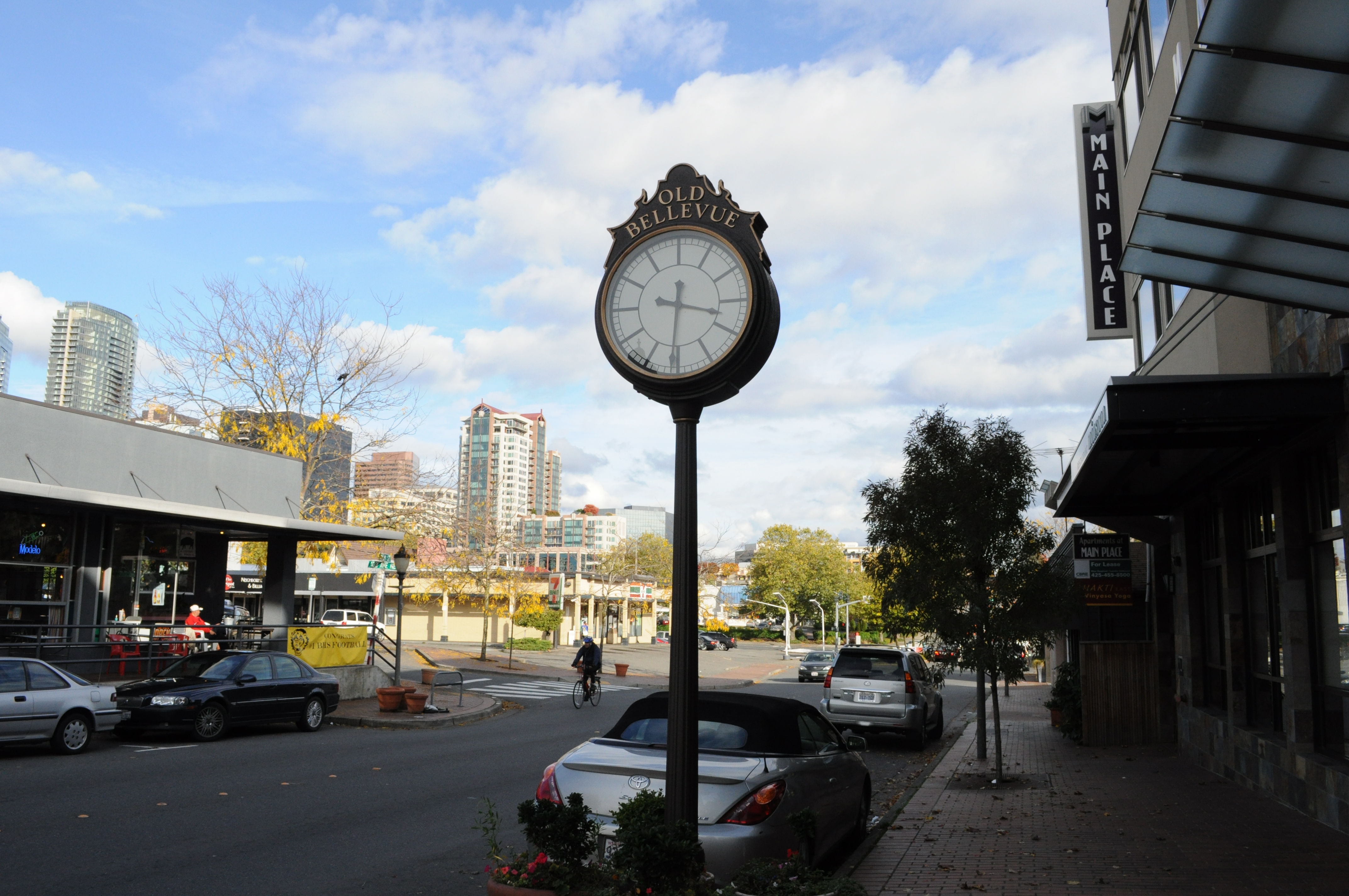 Old Bellevue street clock on a pole with storefronts and high-rise buildings behind it.