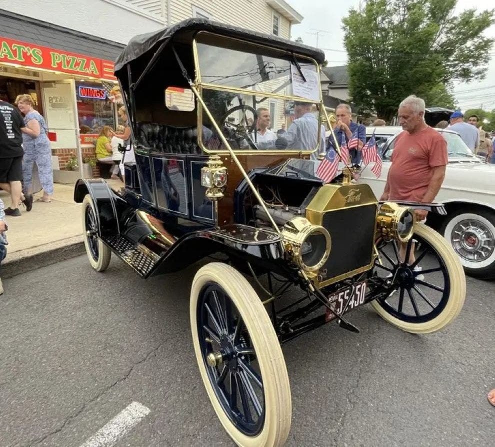 A black antique car with white-wall tires and small American flags on display at an outdoor car show.