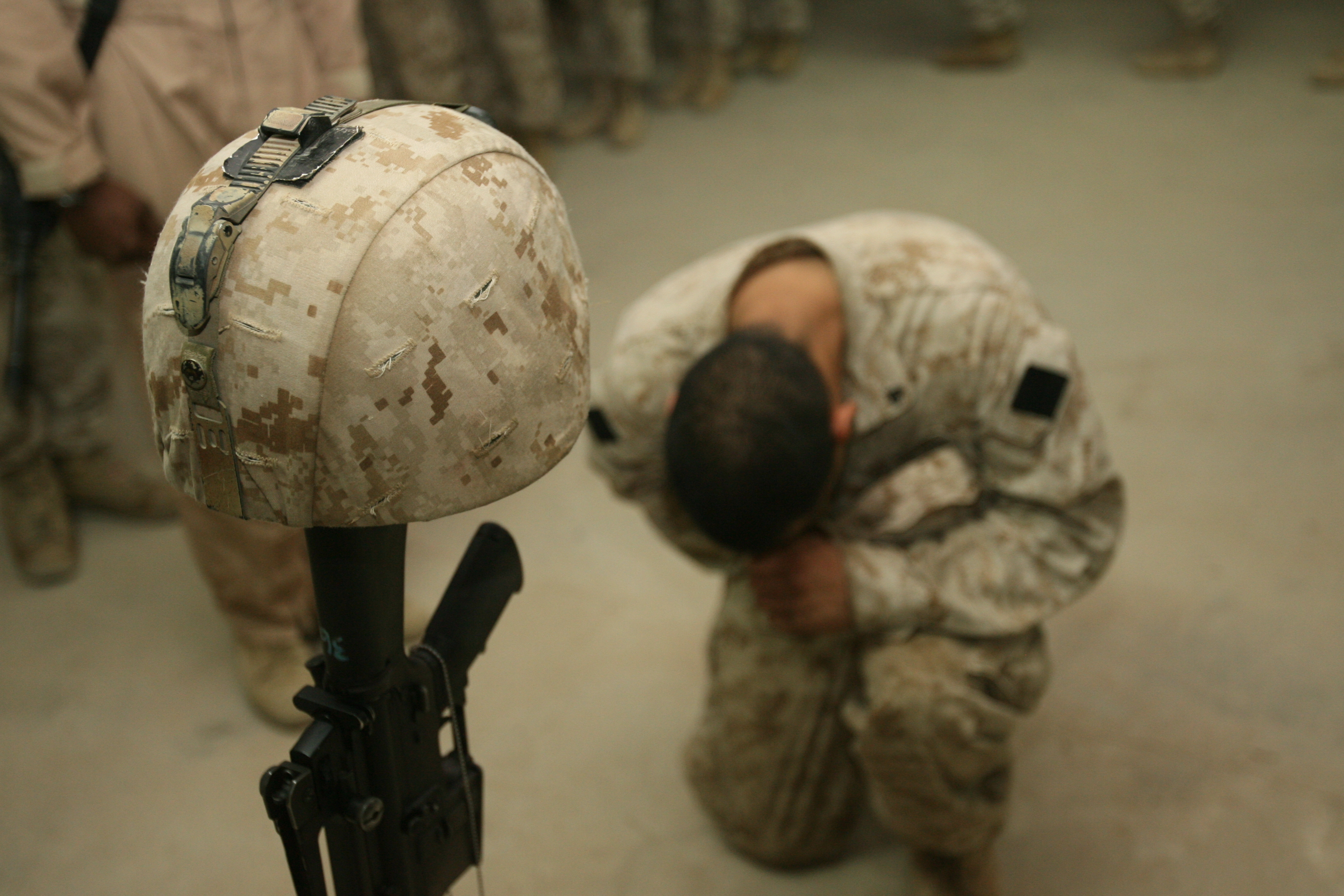 A Marine in camouflage kneels before a battlefield cross memorial indoors.