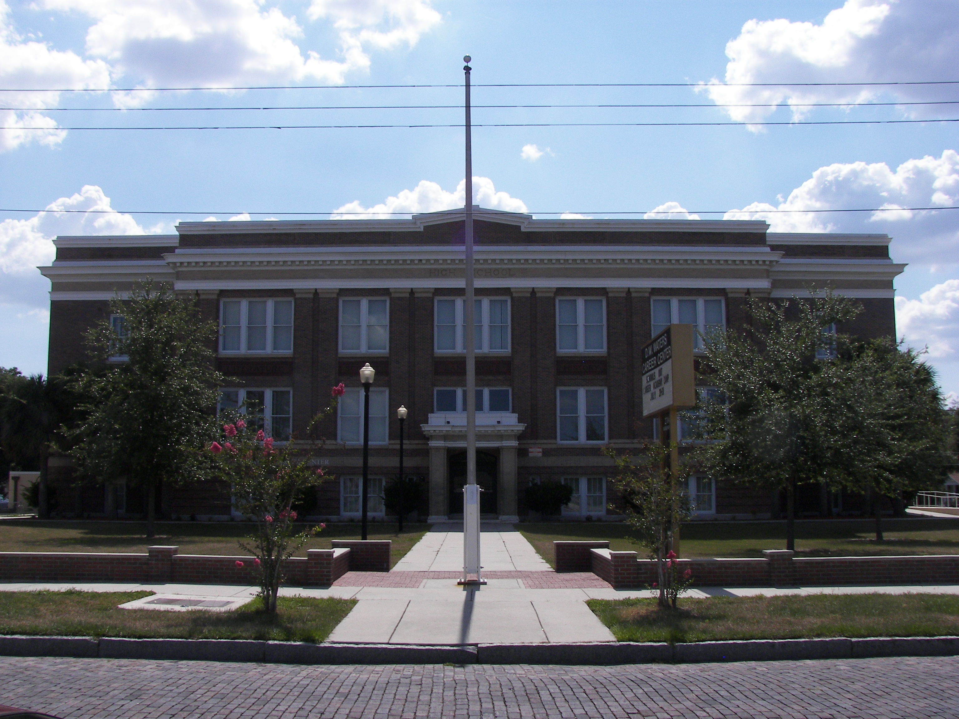 Three-story brick school building with a flagpole and tree-lined walkway.
