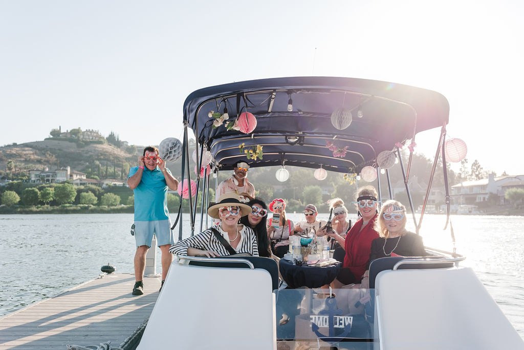 Group of adults in festive attire gathered on a decorated motorboat on a calm lake.