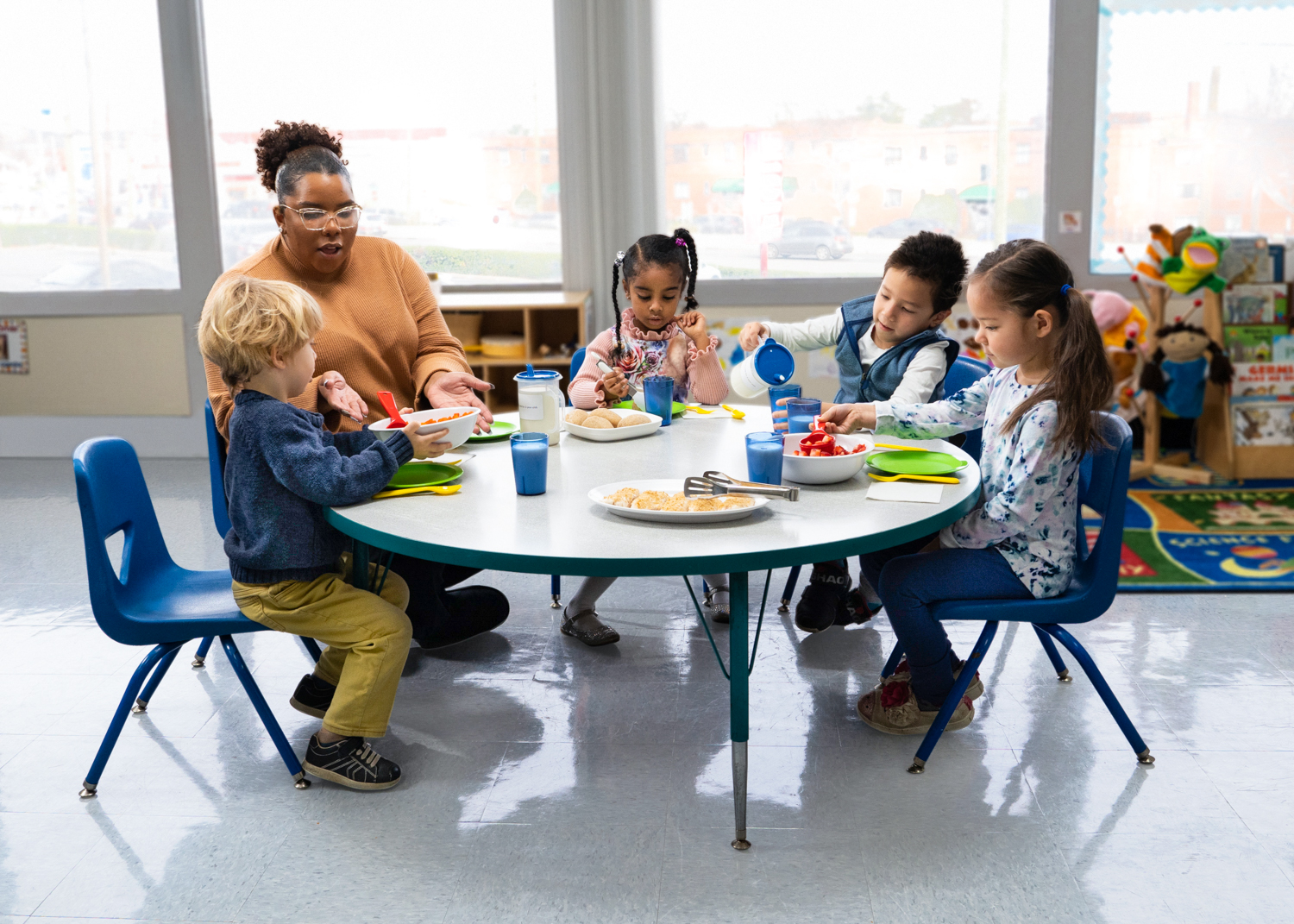 An adult and four young children share a meal at a round table in a preschool classroom.