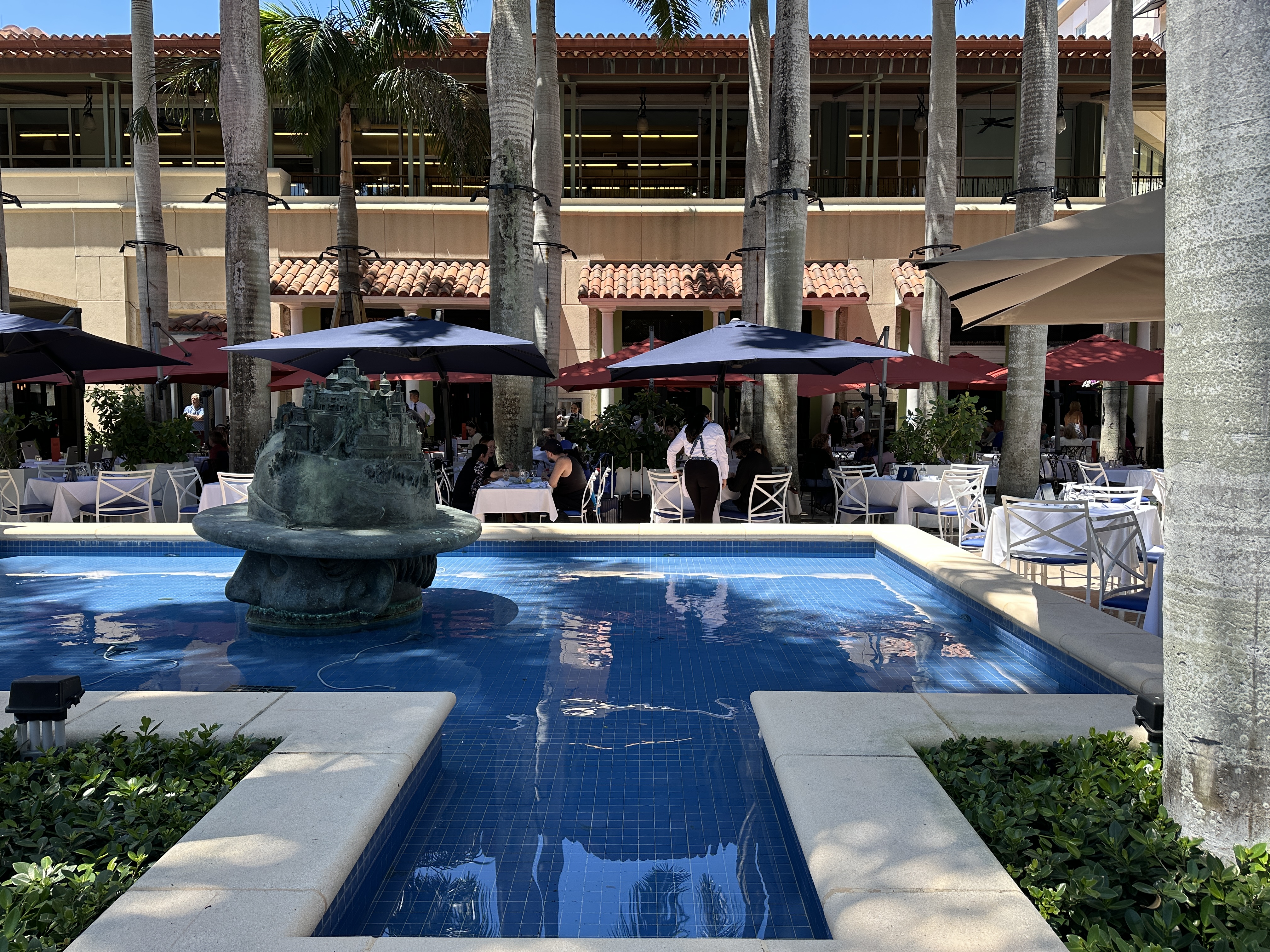 Outdoor courtyard with a bronze fountain, palm trees, and dining tables with purple umbrellas.