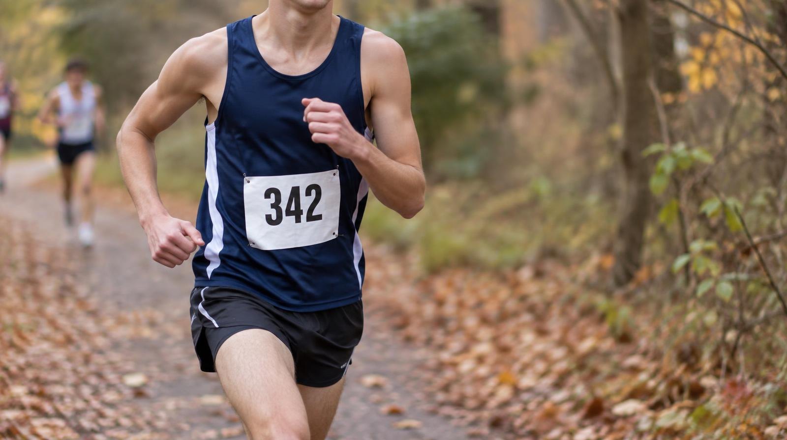 A runner in a racing singlet strides along a leaf-covered wooded trail in autumn.