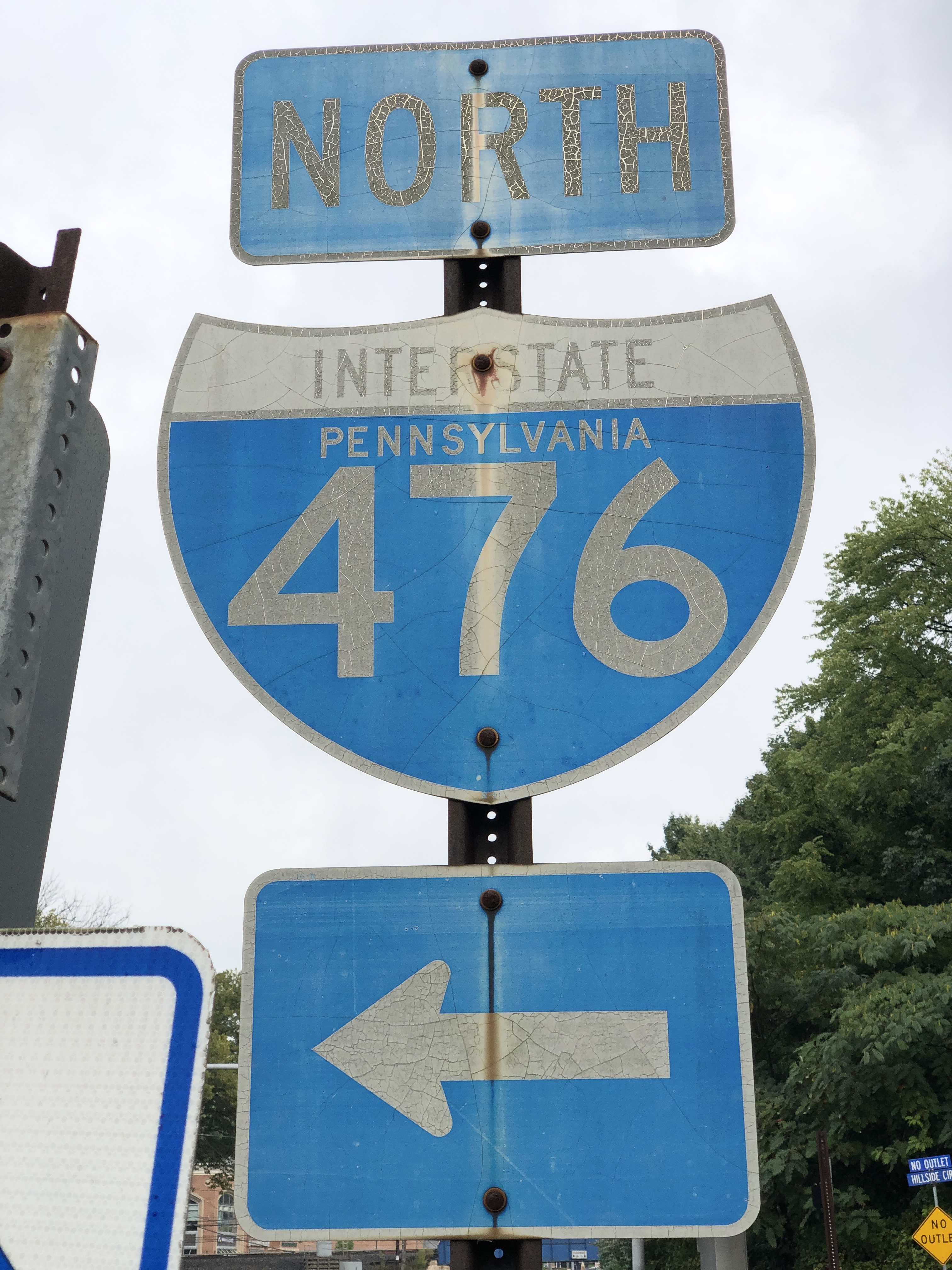 Weathered I-476 North highway sign on a pole along a tree-lined road.