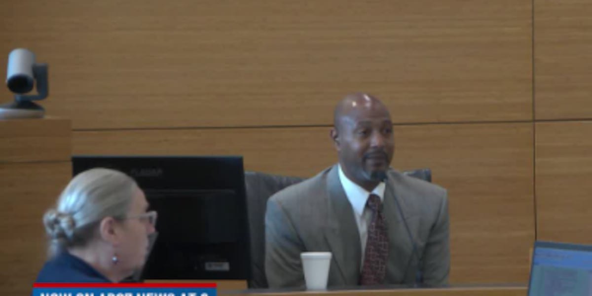 Man in a tan suit seated at a table in a courtroom setting.