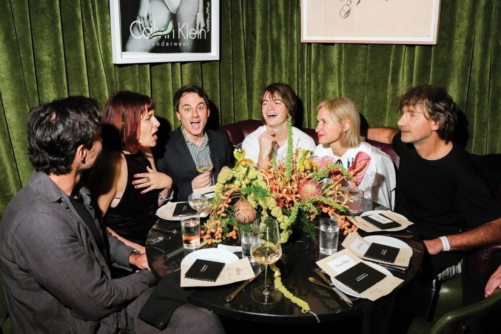 Six people laughing around a round dining table in a dimly lit restaurant with green velvet walls.