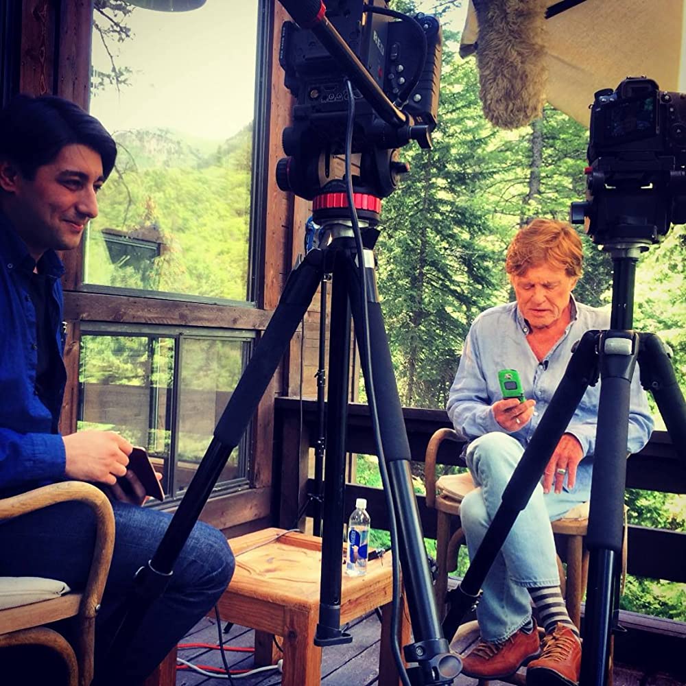 Two men seated outdoors on a deck with camera tripods between them and forested mountains behind.
