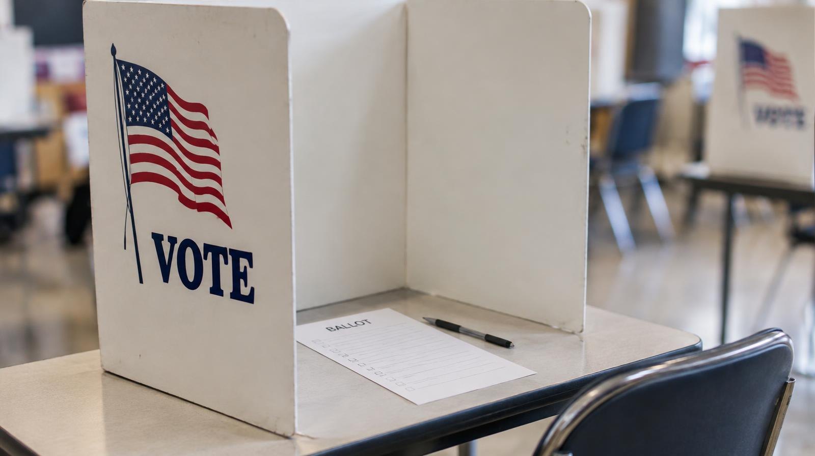 A privacy voting booth with a ballot and pen on a table under fluorescent lighting.