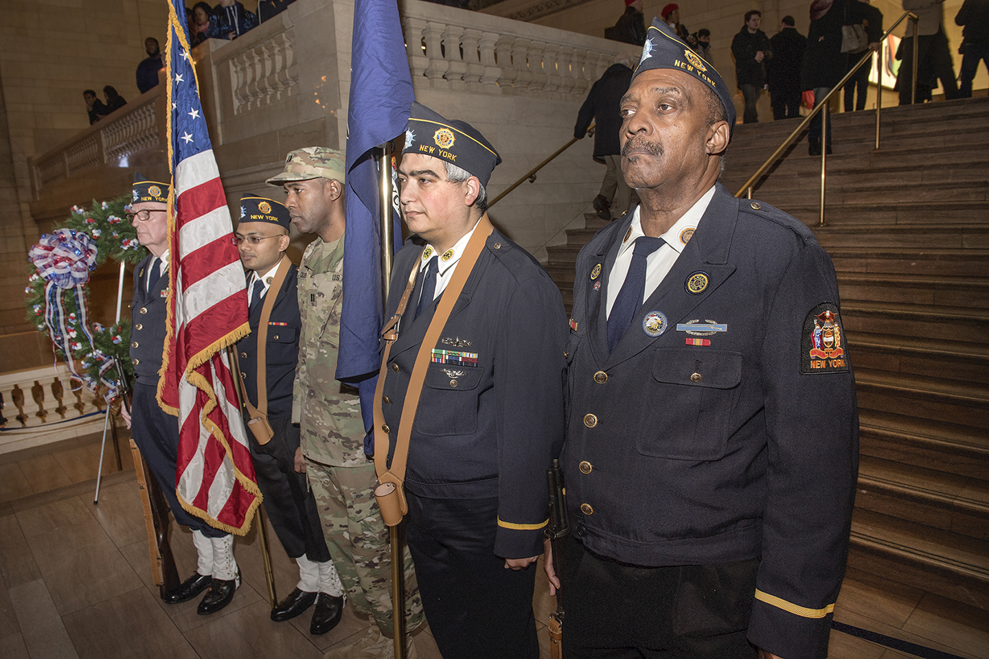 Uniformed American Legion Color Guard members carrying flags at an indoor ceremony.