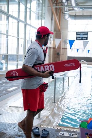 Lifeguard in red cap and gray shirt holds a red rescue buoy at an indoor pool.