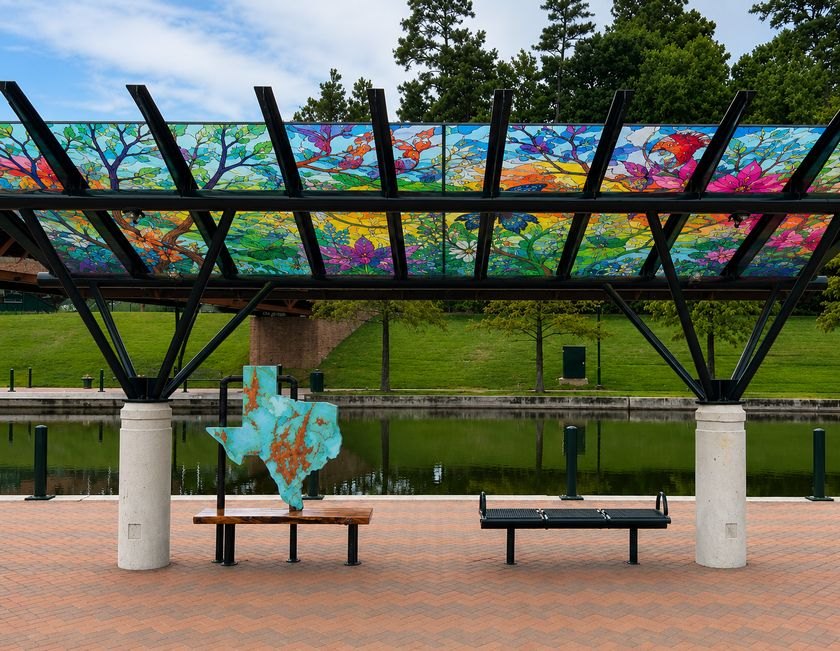 Trolley shelter with colorful tree-patterned glass panels over benches along a waterway.