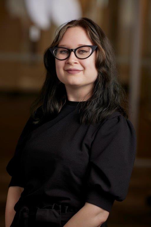 A young woman with long dark hair and black-framed glasses smiling in a professional portrait.