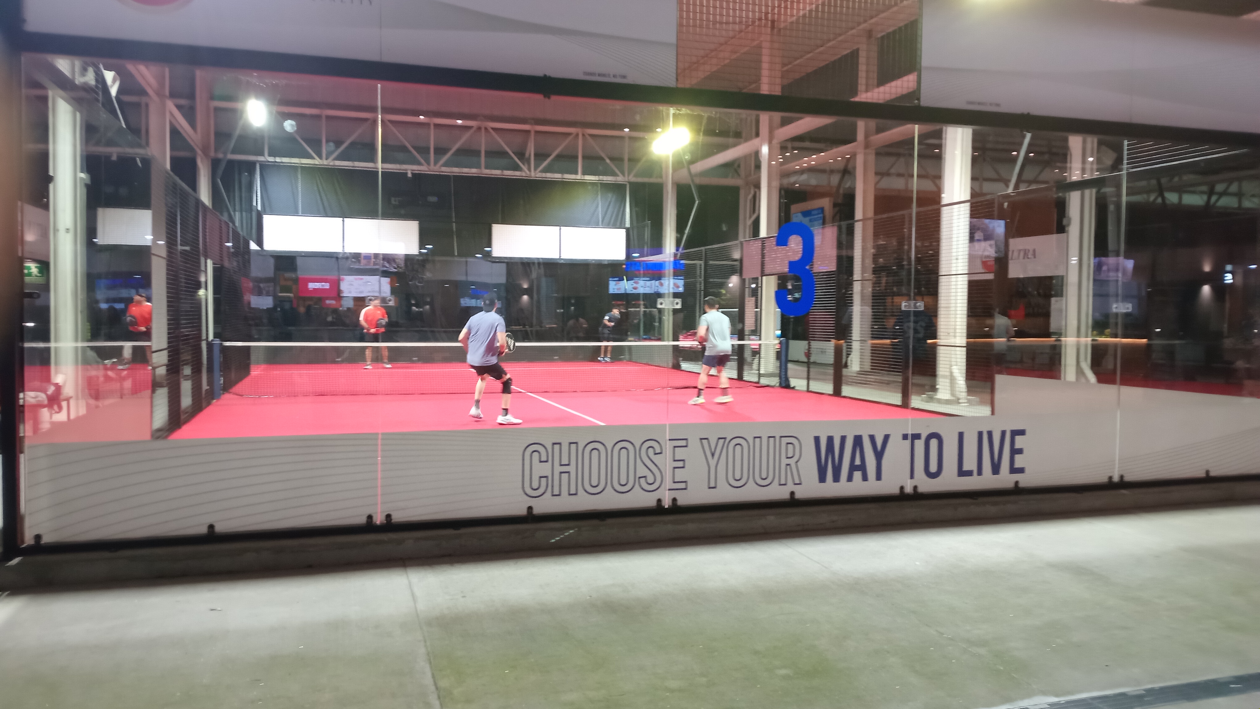 Players mid-game on an indoor padel court with red turf and glass walls.