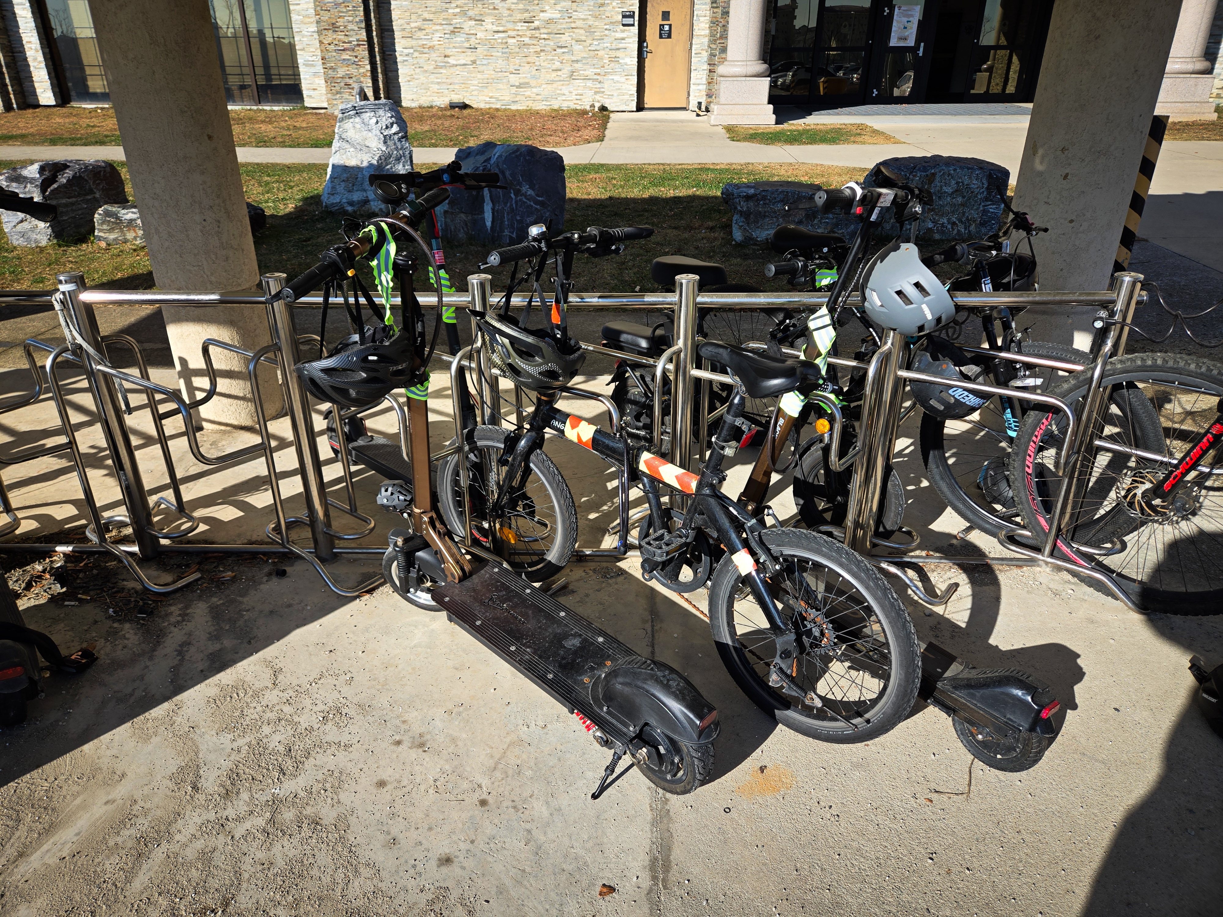 E-bikes, electric scooters, and bicycles parked together at an outdoor metal bike rack.