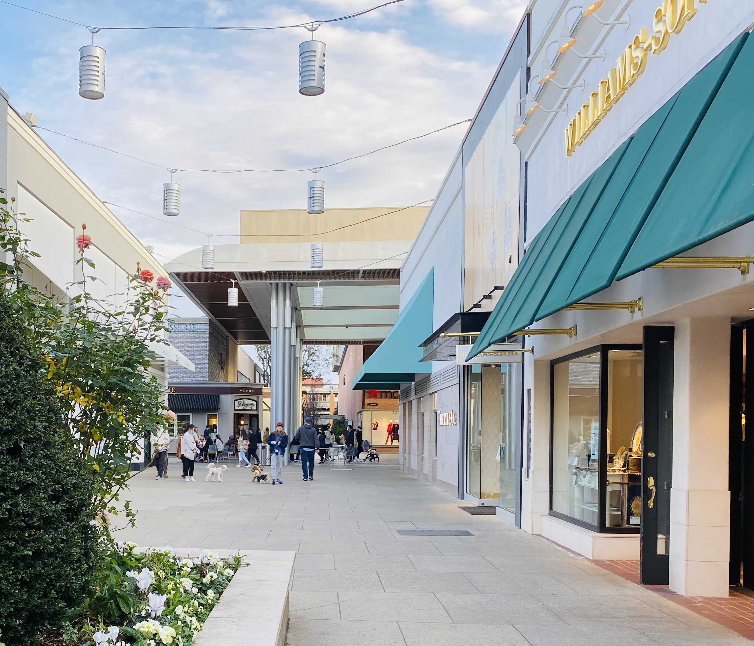 Shoppers walk along an outdoor mall corridor lined with storefronts and hanging lights.