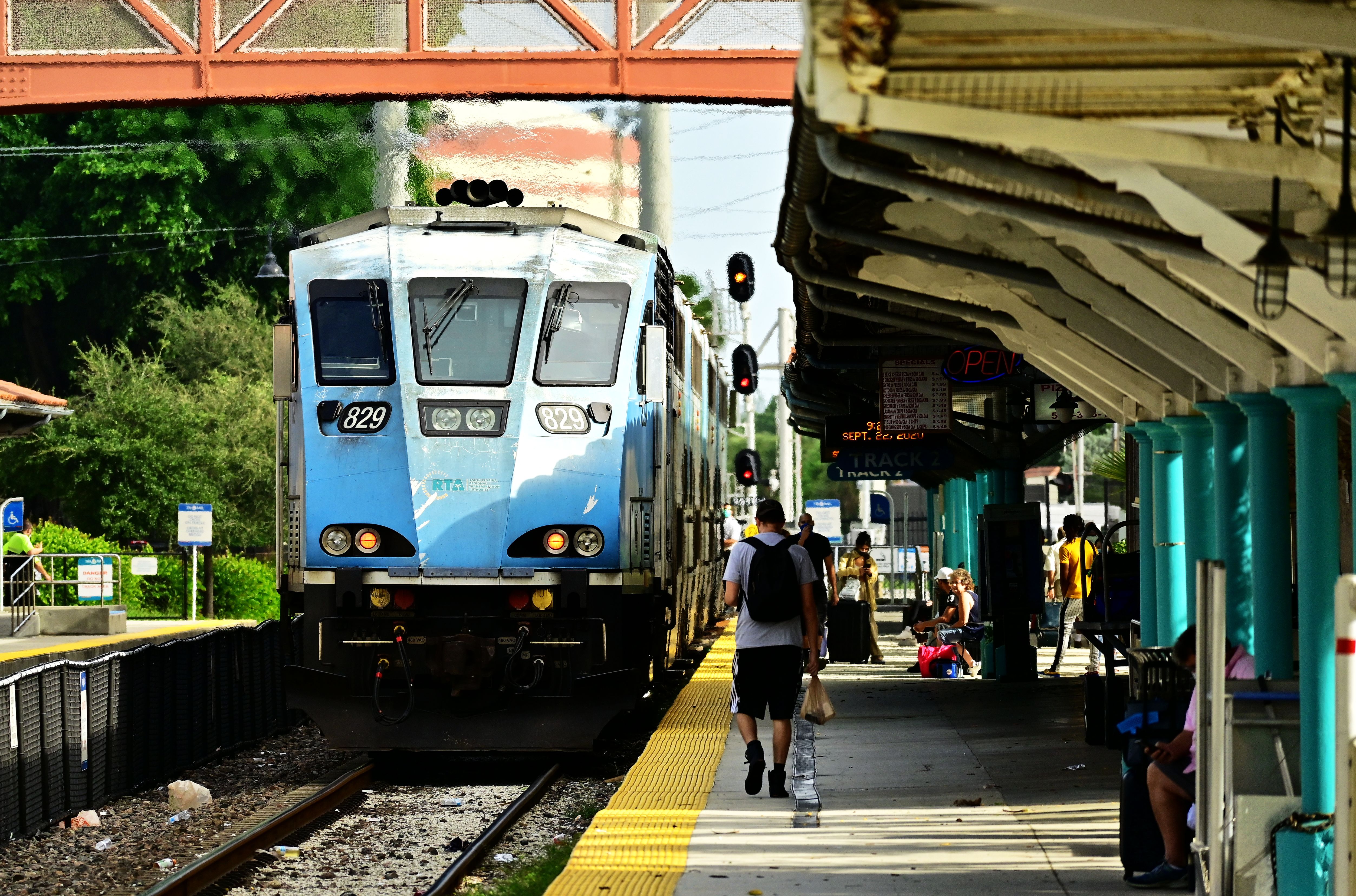 A light-blue Tri-Rail commuter train arrives at a covered station platform with waiting passengers.