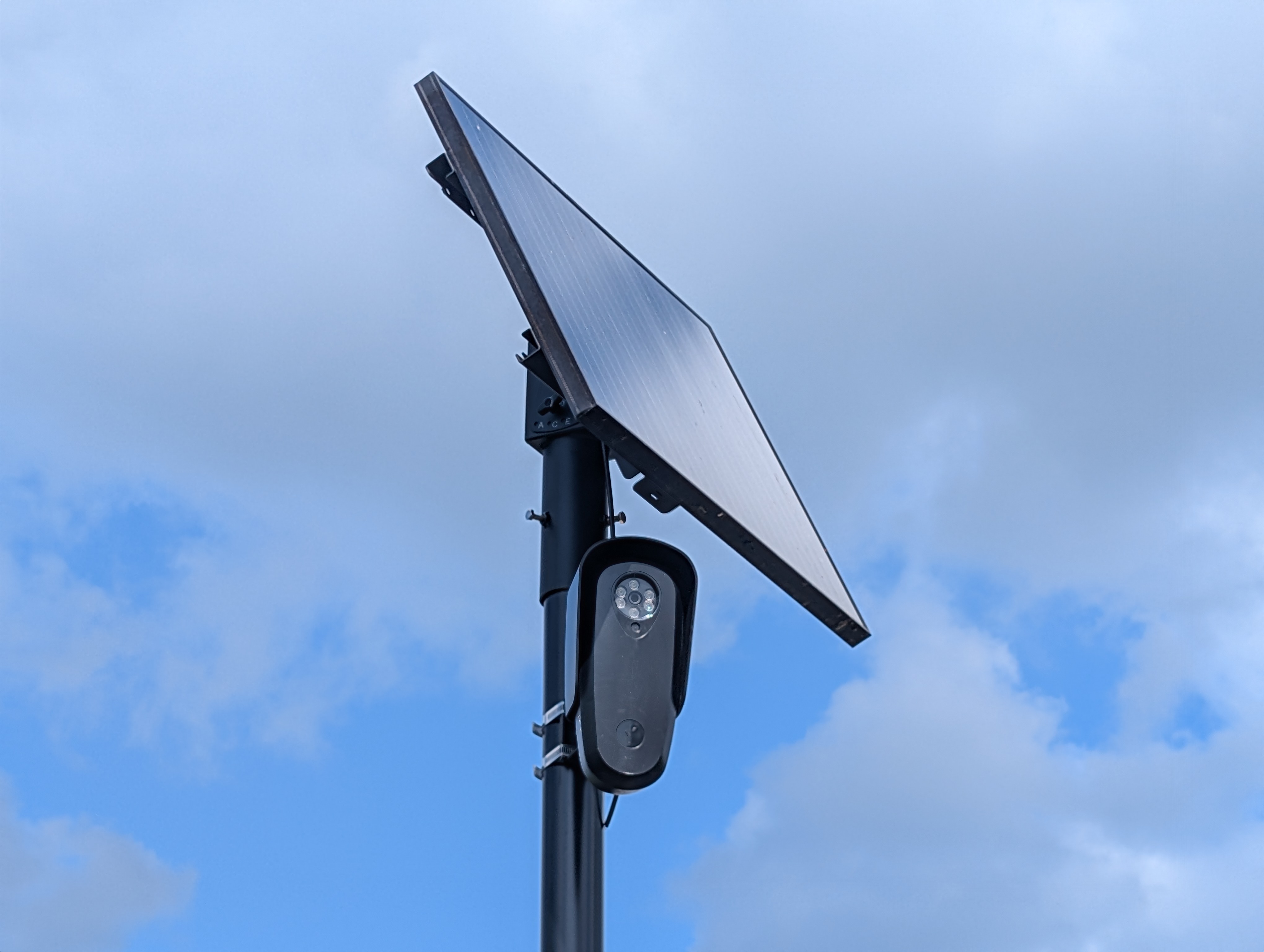 Close-up of a black Flock Safety license plate reader camera on a pole against a blue sky.