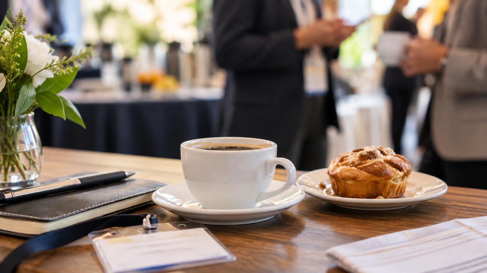 A coffee cup and pastry on a table with name badges and notebooks nearby.