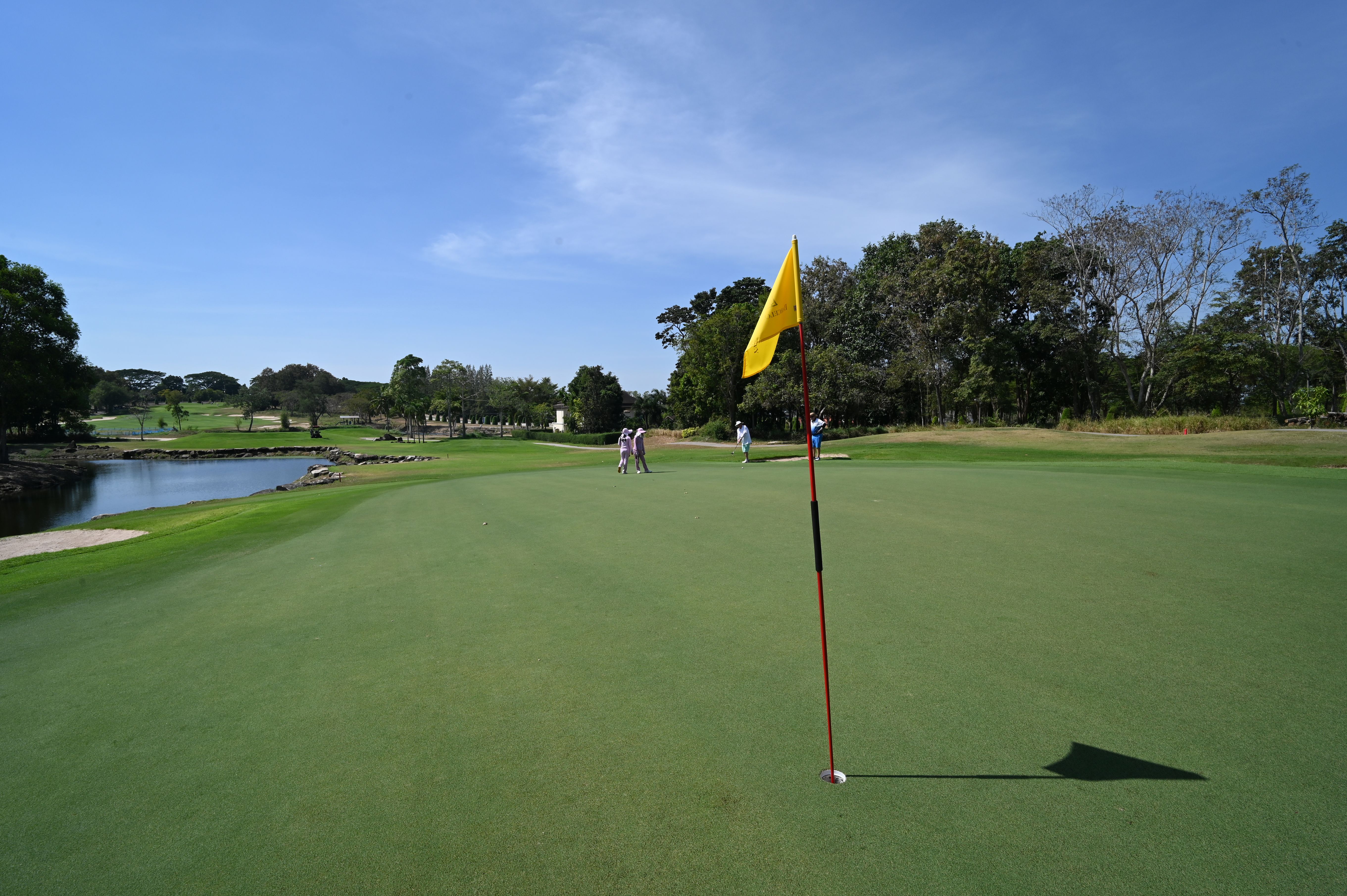 Golfers walk toward a putting green with a yellow flag on a sunny course.