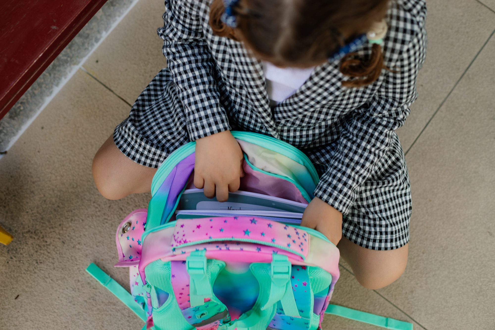Directly above view of small girl with brown hair looking for something in colorful backpack