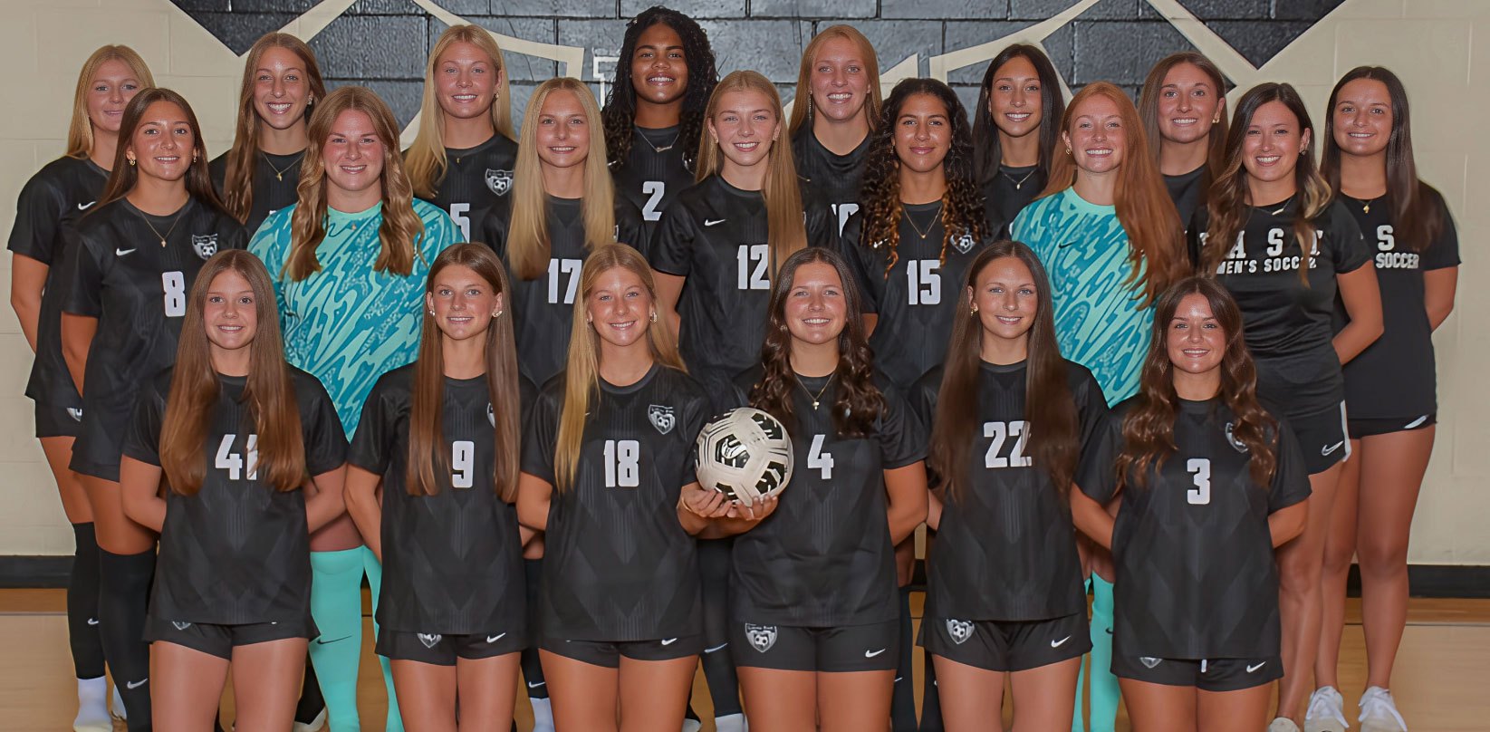 About 20 young women in dark soccer uniforms pose in three rows for a team photo indoors.