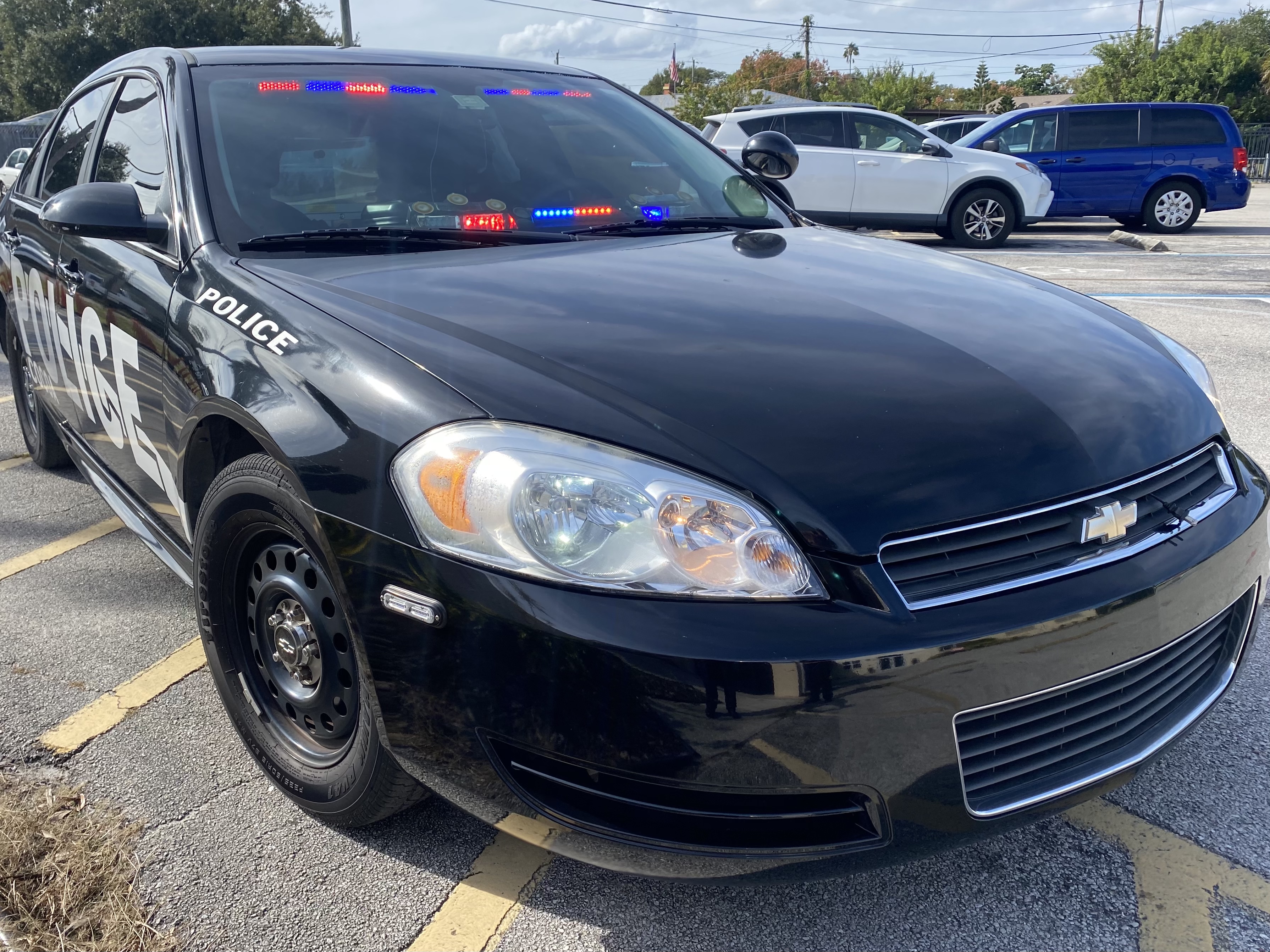 Black police patrol car with red and blue emergency lights activated, parked in a lot.