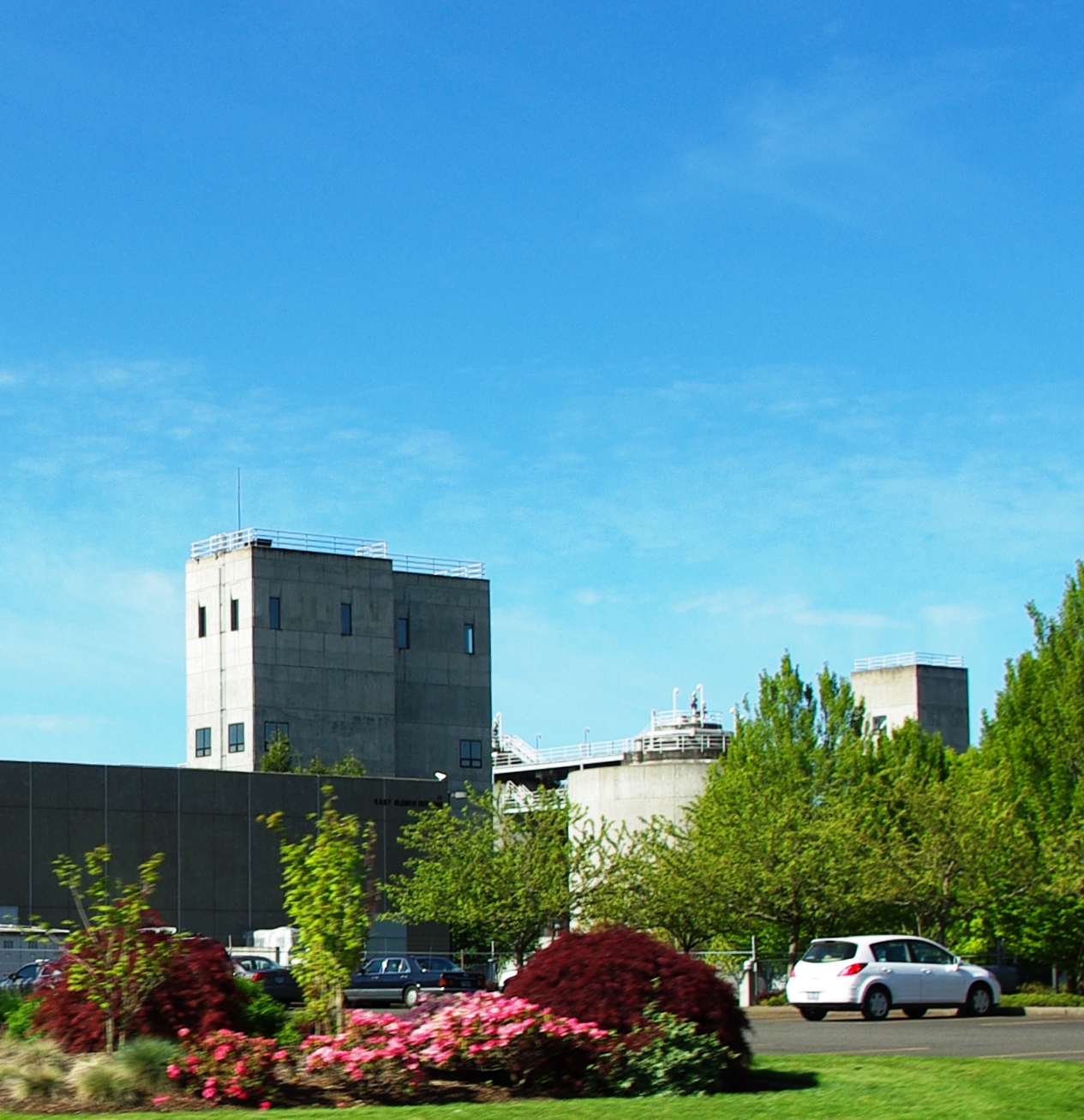 Concrete towers of a water treatment plant rise above flowering shrubs on a sunny day.