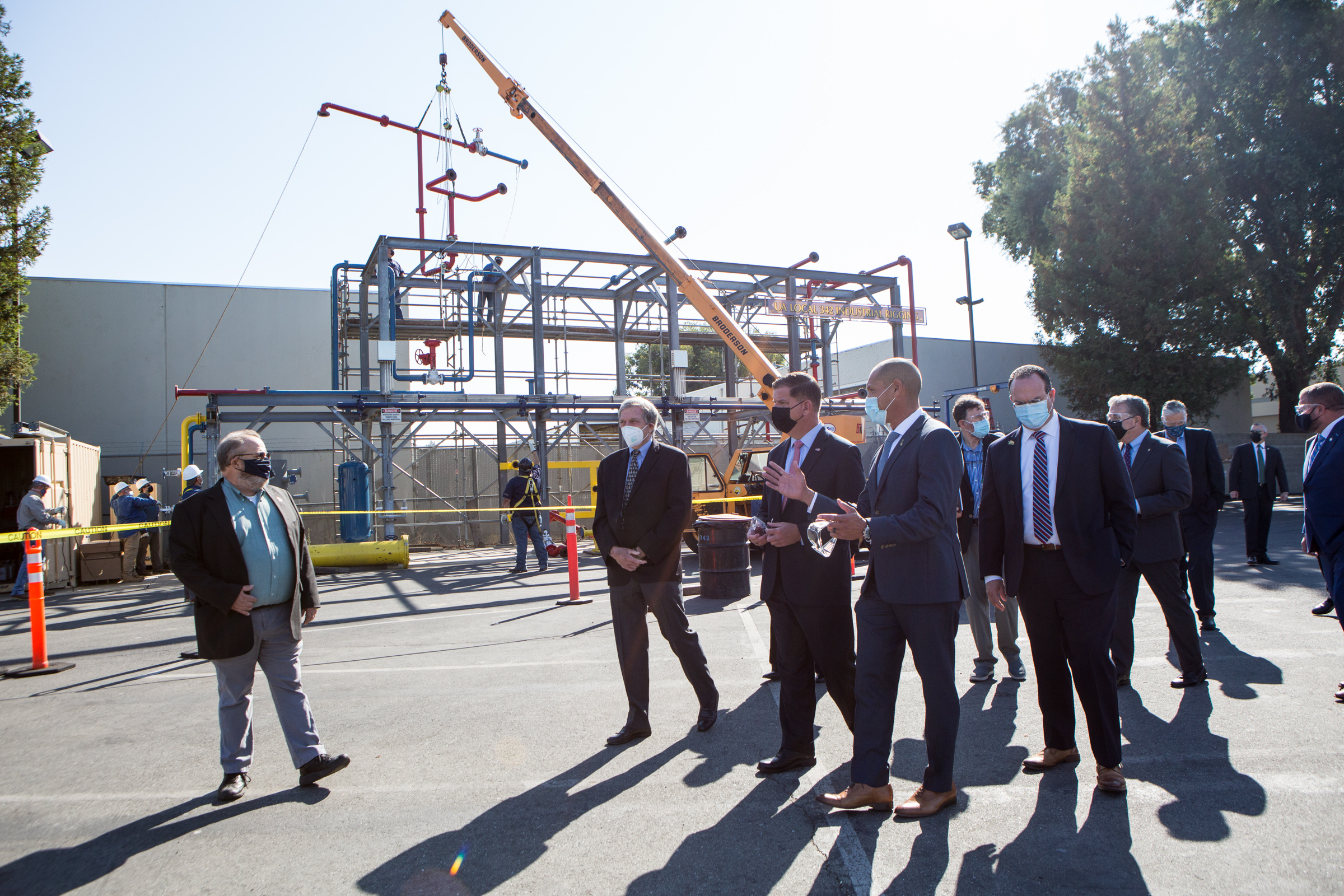 Masked officials in suits walk outdoors at an industrial training facility with a crane in the background.