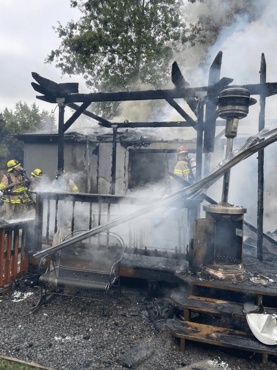Firefighters in full gear spraying water on the heavily charred frame of a burned house with smoke rising.