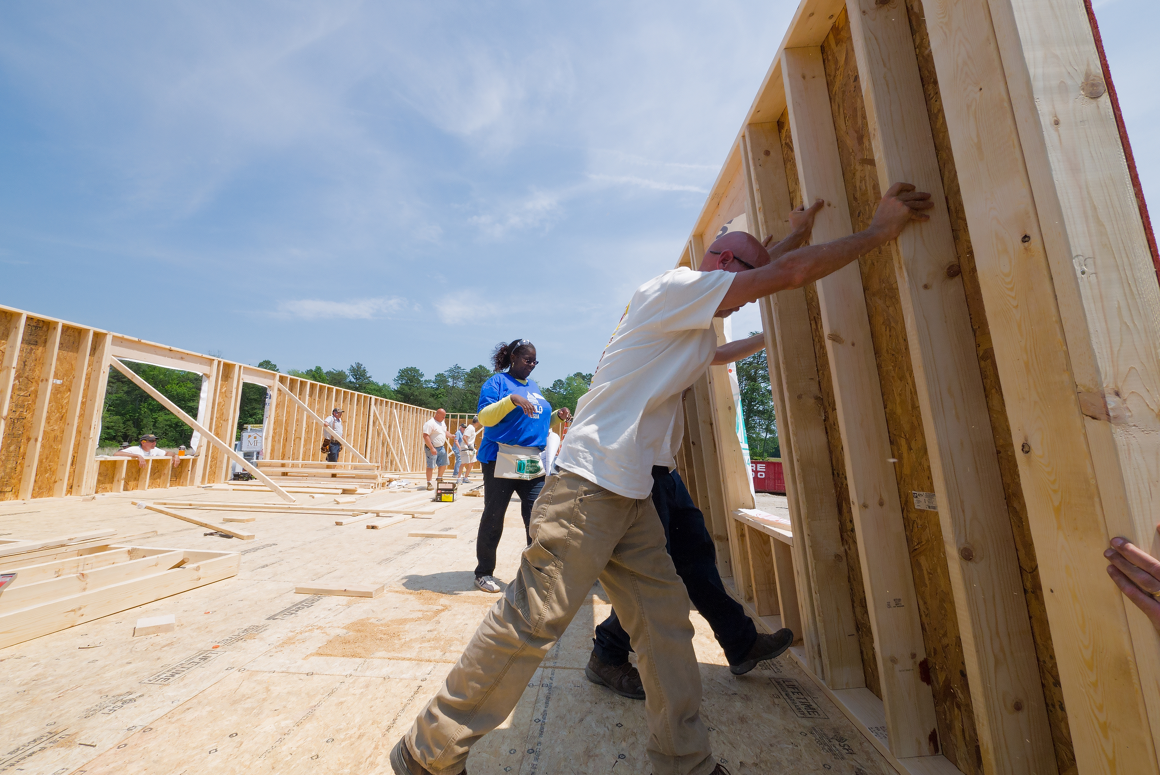 Construction workers raising a wooden framed wall section at a sunny residential build site.
