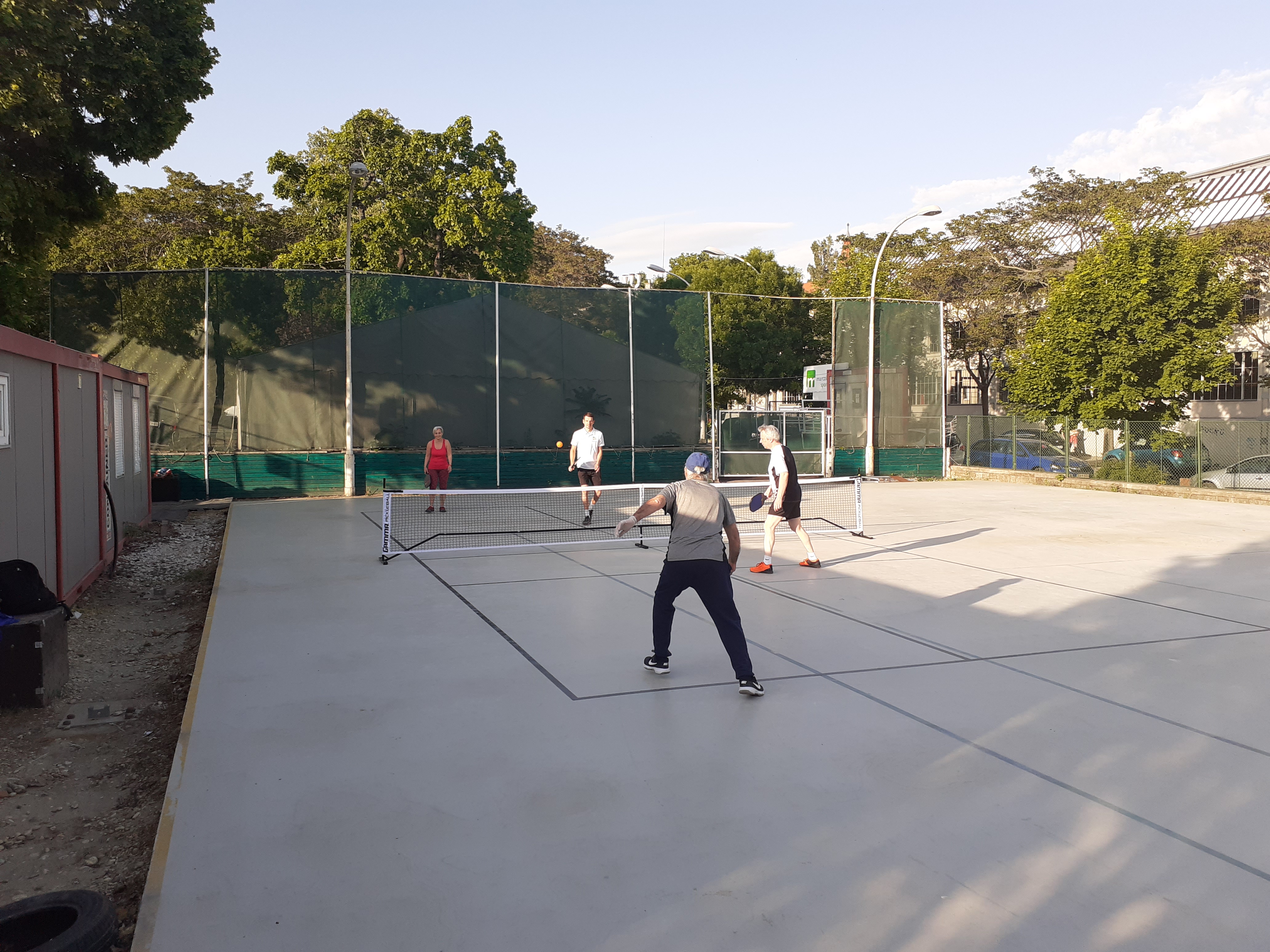Four players on an outdoor pickleball court with a low net and green windscreen fencing.