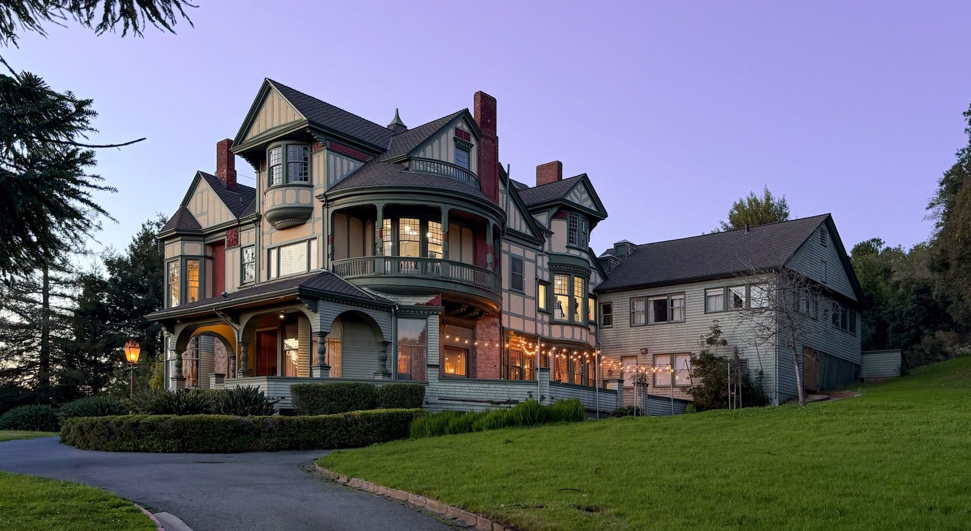 A Victorian mansion lit from within at dusk, with string lights along a garden path.