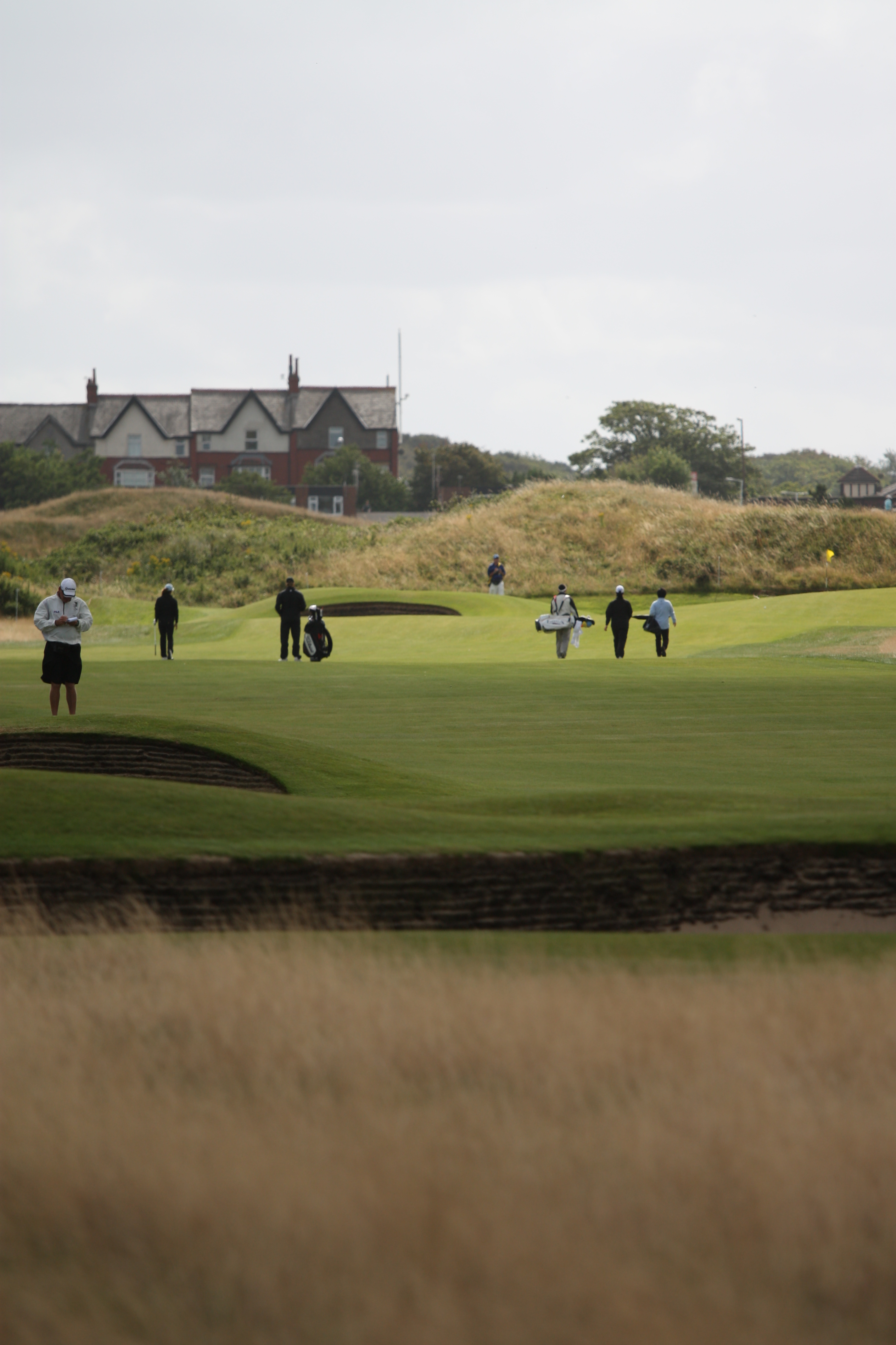 Golfers and caddies walk along a fairway with sand bunkers on either side.
