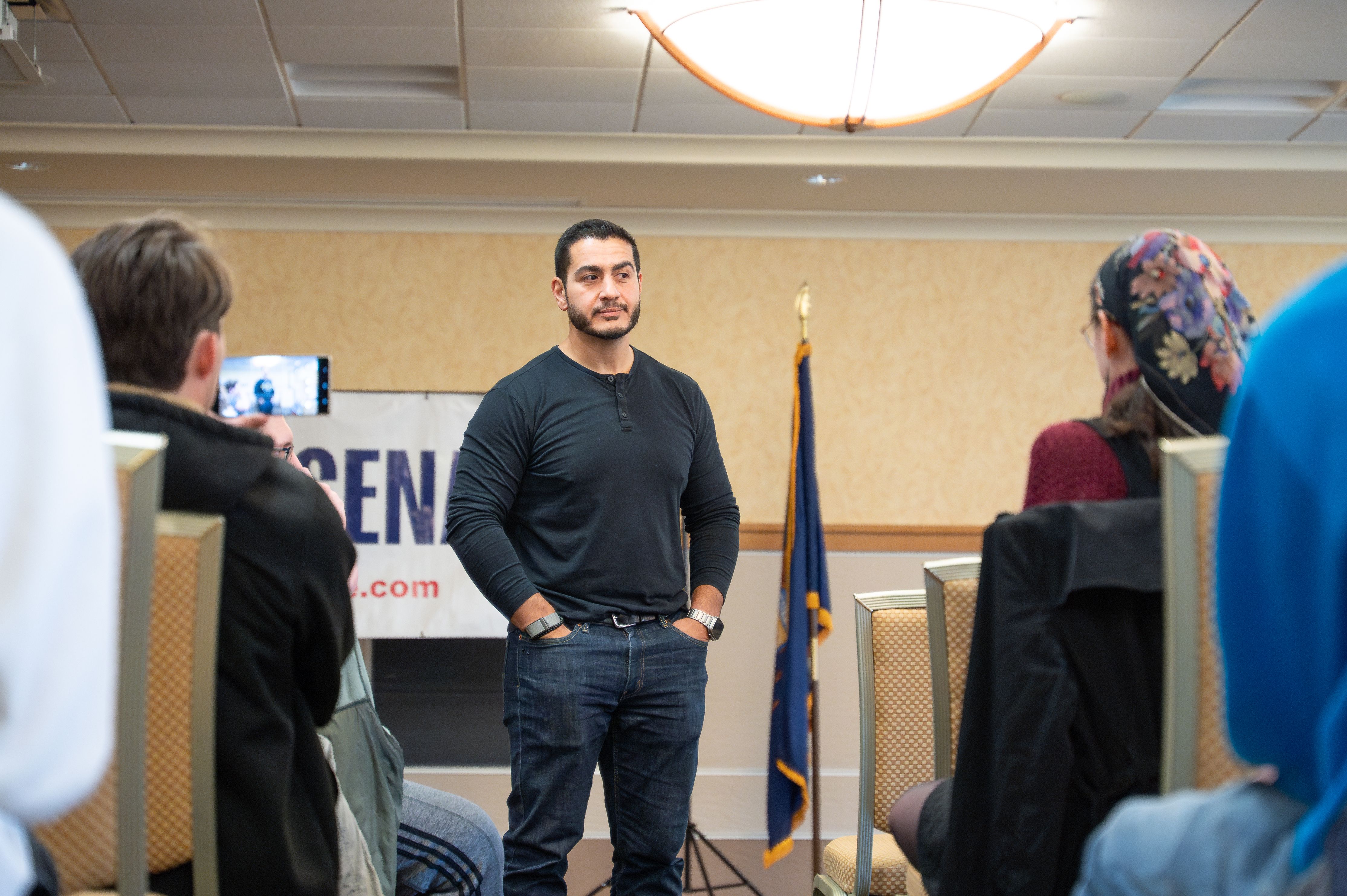 A man in a black shirt speaks to a small seated audience in a conference room with a state flag behind him.