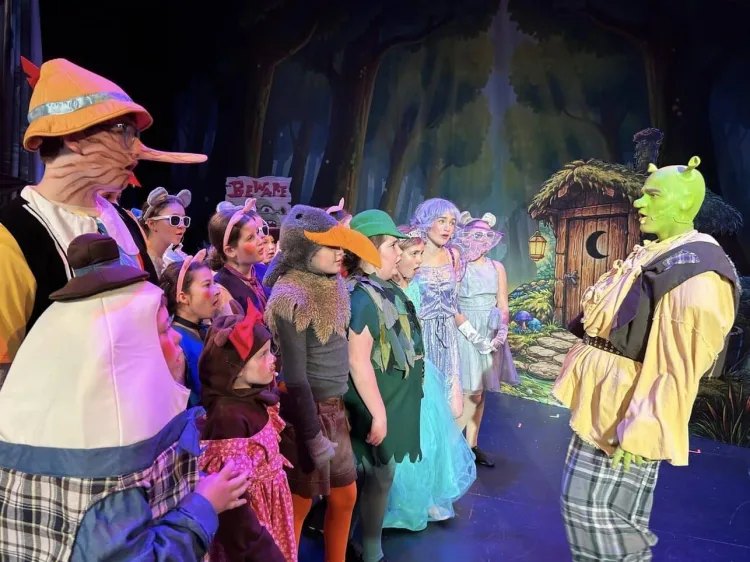 Young performers in theatrical costumes on a stage set, including a Shrek character in green makeup.