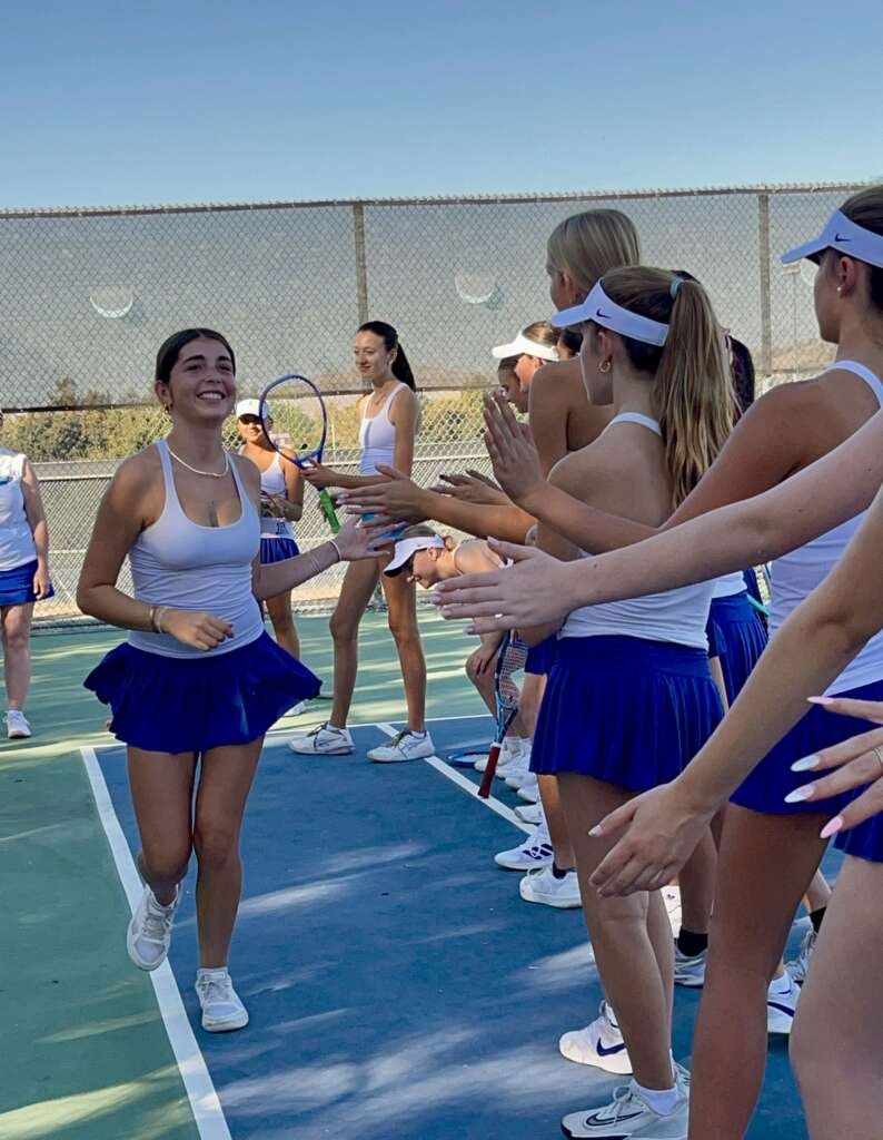 Girls tennis players in white tops and blue skirts form a high-five line on an outdoor court.