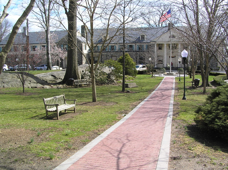 Brick pathway lined with bare trees and benches leading to a large institutional building with an American flag.