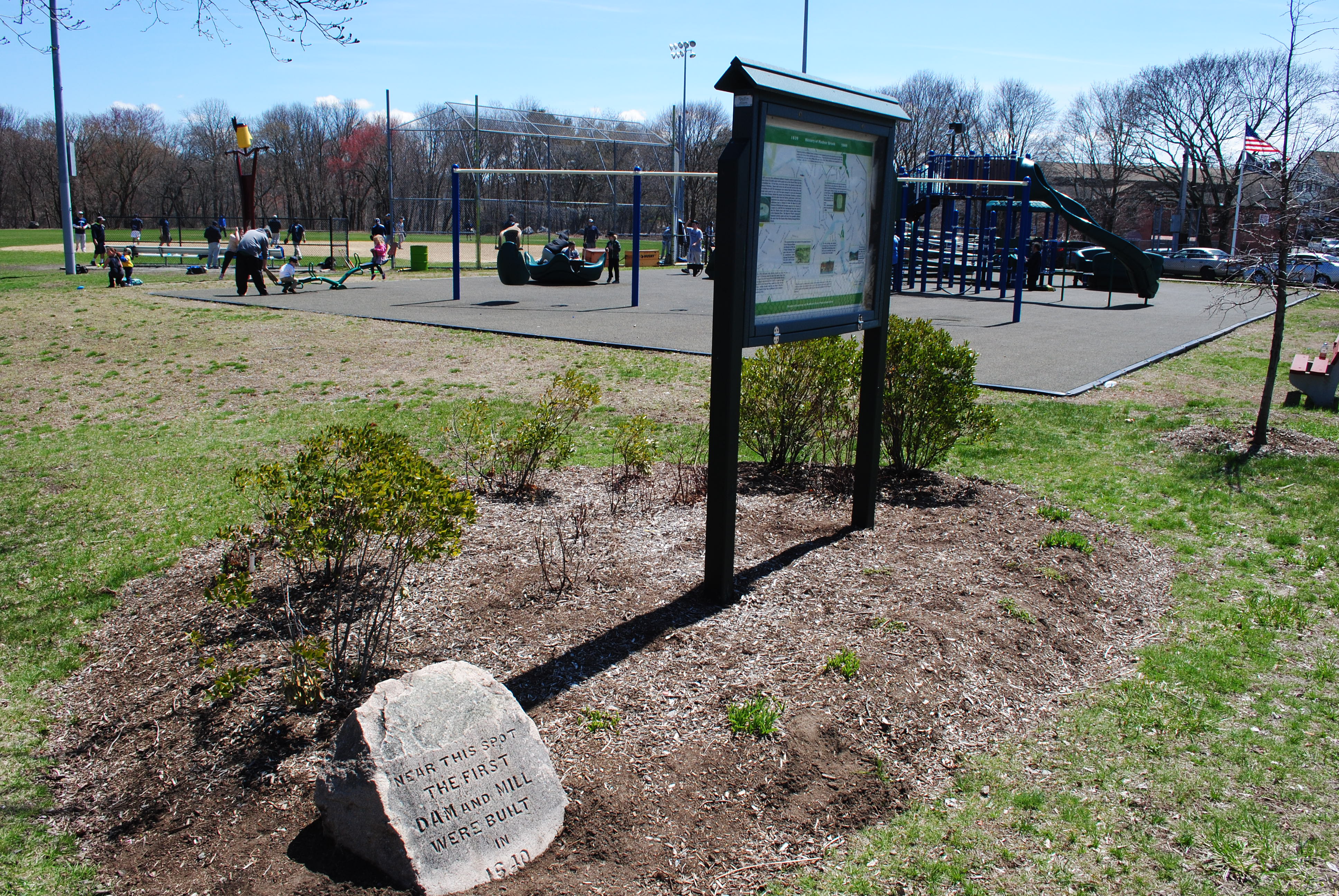 A park with a stone historical marker, playground equipment, and a ball field under blue sky.