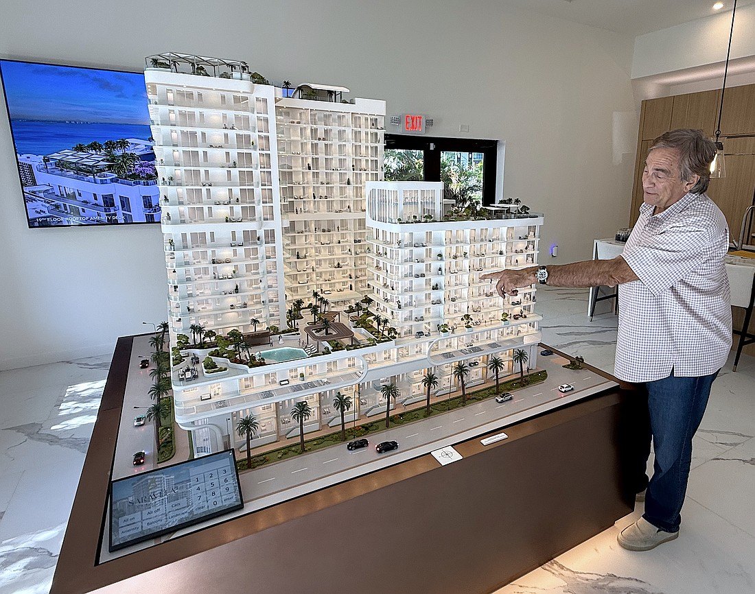 Man in plaid shirt points at a detailed scale model of two high-rise towers with a pool courtyard.