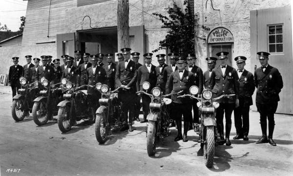 Black-and-white photo of uniformed officers with motorcycles outside a police building.