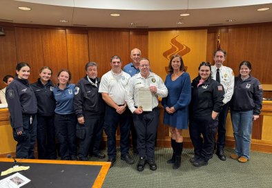 Group of people in EMS uniforms standing in a wood-paneled council chamber, one holding a proclamation.
