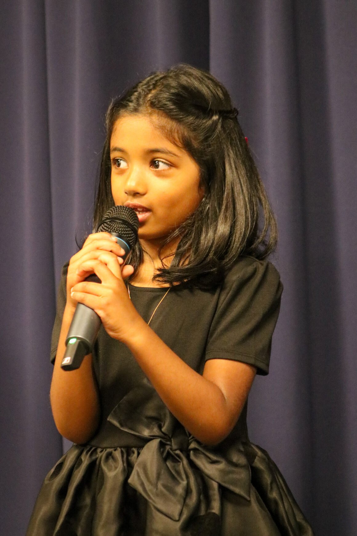 A young girl in a black dress sings into a microphone on a darkened stage.