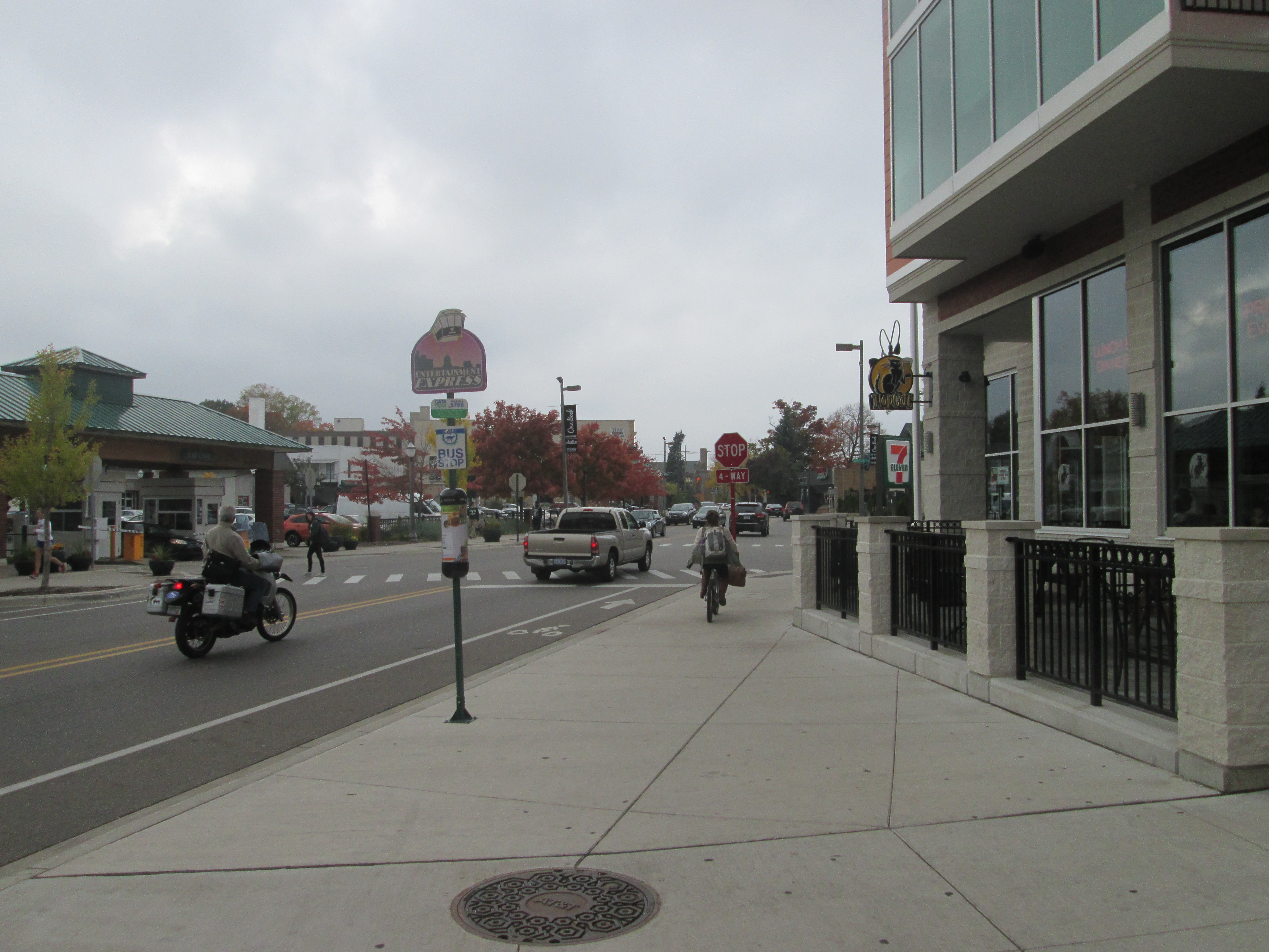 Daytime street view of East Lansing with storefronts, pedestrians, and passing vehicles.