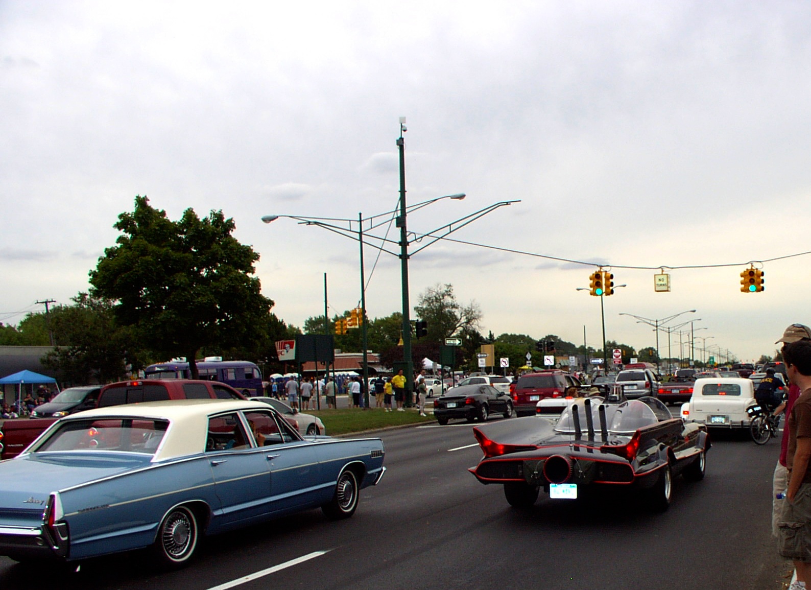 Classic cars including a replica Batmobile cruising a busy urban boulevard at a traffic light.