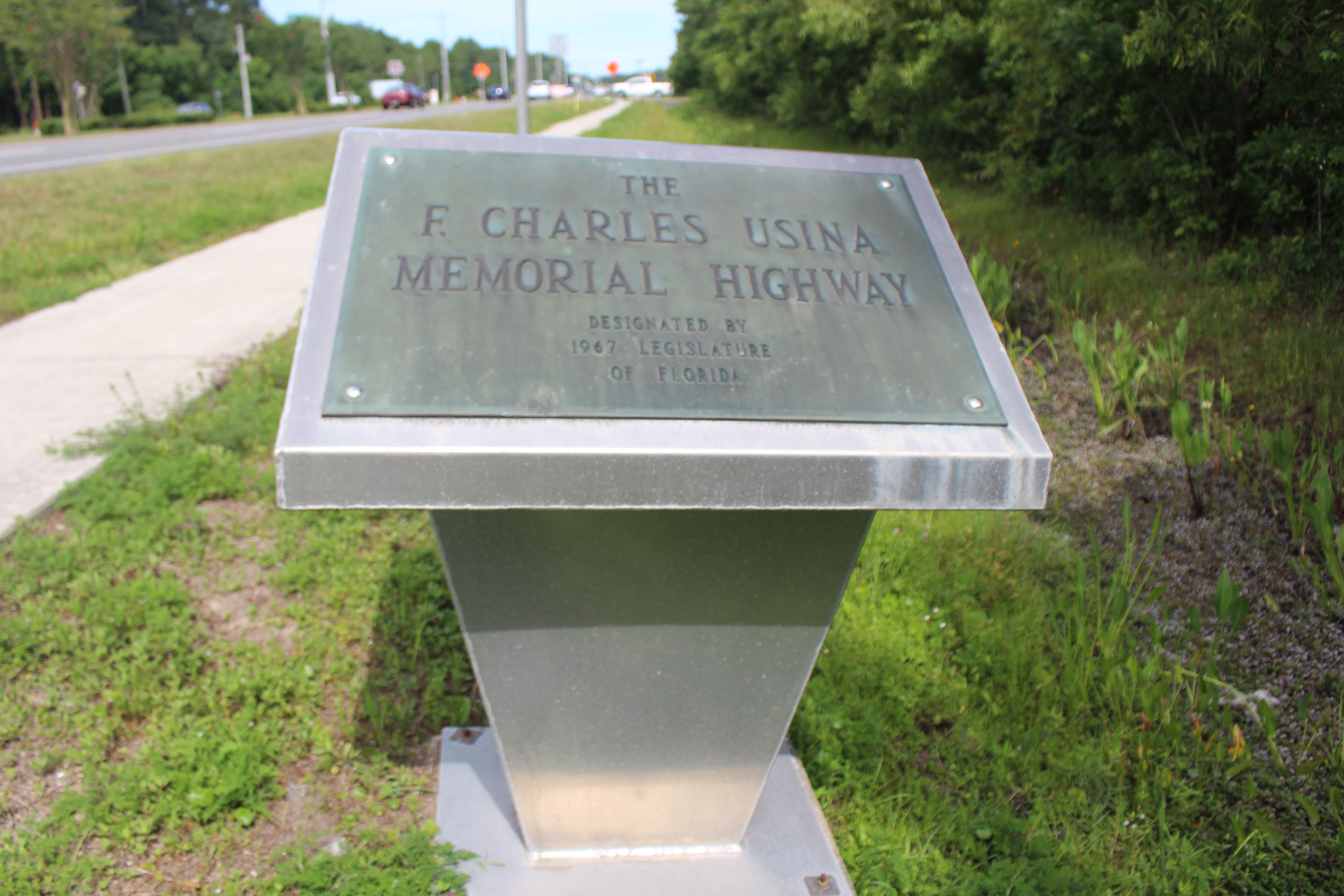 Bronze plaque on a metal pedestal along a roadside, with trees and traffic in the background.